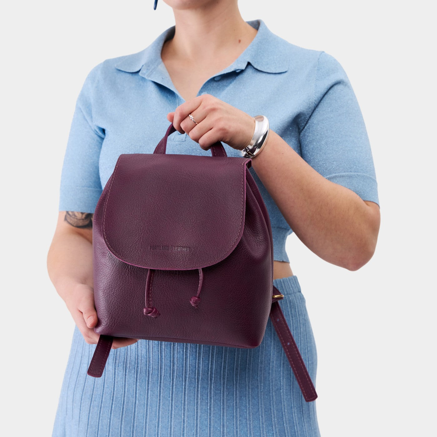 Plum | Pebbled purple leather drawstring backpack being held by a person wearing a blue sweater.