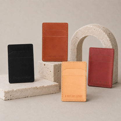 Naturale | Four slim leather card holders in black, tan, brown, and rdark brown, arranged on and around light stone blocks and an arch against a warm beige studio background.
