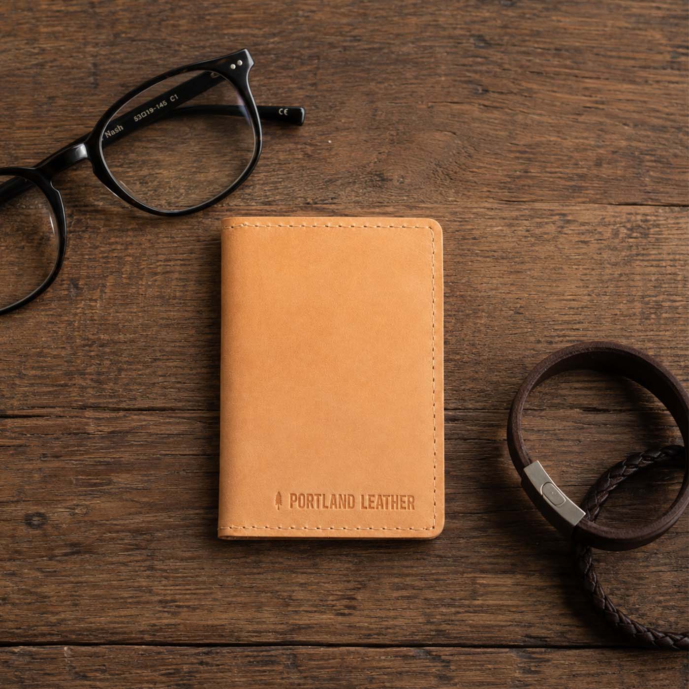 Naturale | A leather card holder styled on a rustic wooden tabletop with black eyeglasses and a dark leather bracelets
